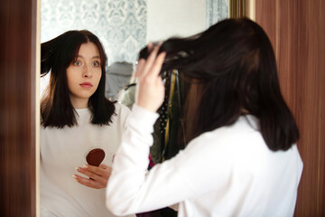 Girl teenager in surprise looks at her hair in the reflection of the mirror, seasonal problem of hair loss, the effects of stress, improper hair care