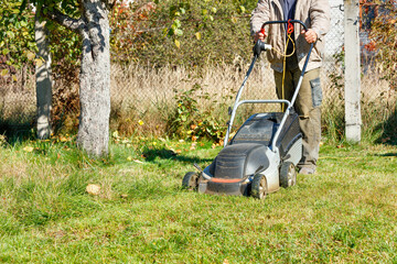 A gardener mows green grass with an electric lawn mower on his backyard on a bright sunny autumn day.
