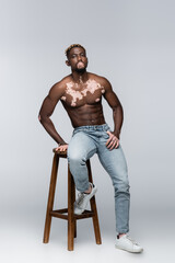 shirtless african american man with vitiligo and stylish hairstyle posing on high stool on grey.