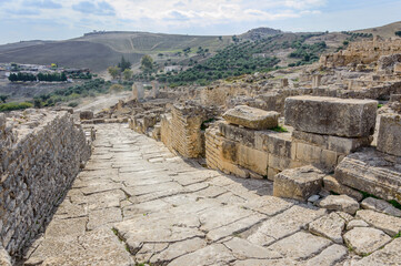 Ruined street of the antique Roman city of Thugga in Tunisia, Africa. Blue sky, paved road, sunlight and shadows on the ancient stone walls, hills with green olive trees on the horizon
