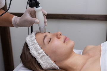 young woman in beauty center performing radio frequency therapy of facial complexion for skin...