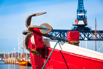 Vintage Maritime Harbor Details / Rusty anchor on bow of old wooden cutter boat at city port background with huge gantry crane (copy space) - 480469704