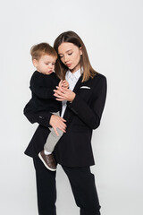 stylish mother in suit holding in arms toddler son isolated on grey.