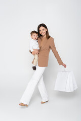 joyful mother holding in arms toddler son and shopping bags while walking on grey.