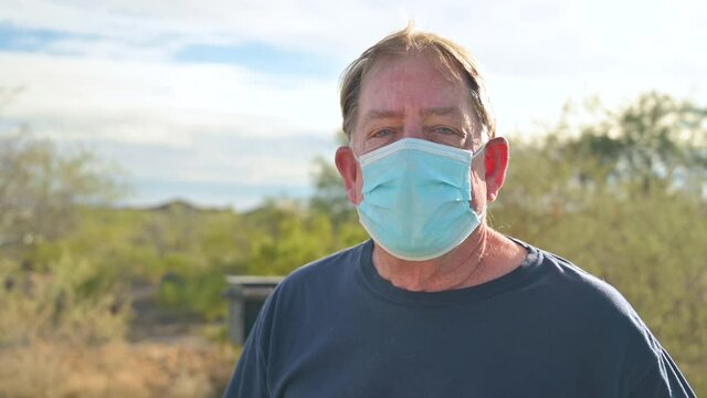 Senior male outdoors in nature, wears face mask during Coronavirus Covid19 pandemic and Delta and Omicron variant spread. Looks at camera.