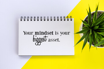 Inspirational motivational quote on a notepaper book - Your mindset is your biggest asset. Business flat lay with plant in a small pot on the white and yellow table background. Positive mind concept.