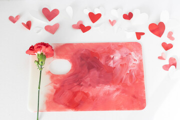 Valentines Day or wedding, invitation, scattered painted small cut out red, pink and white hearts with a red carnation on a colorful paint palette, copy space