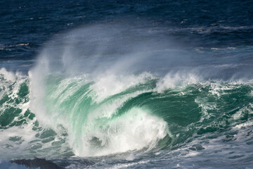 An angry turquoise green color massive rip curl of a wave as it rolls along a beach. The white mist and froth from the wave are foamy and fluffy. The Atlantic Ocean in the background is deep blue. 
