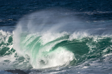 An angry turquoise green color massive rip curl of a wave as it rolls along a beach. The white mist and froth from the wave are foamy and fluffy. The Atlantic Ocean in the background is deep blue. 