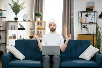 Mindfullness, work at home and skin care. Young bearded man with towel on his head and patches under eyes, sitting on sofa at home with laptop, meditating with eyes closed and hands in zen sign