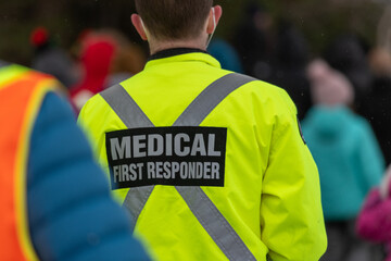 Medical first responders walking along a road wearing black wool stocking caps, yellow reflective coats with the medical first responder in grey letters and across. The EMT is carrying a first aid kit