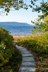 A curved footpath or trail of concrete blocks leading to a sandy beach. The entrance has green and yellow shrubs and trees. There's a mountain in the distance along the riverbank on a sunny day.