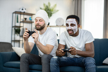 Two young multiethnic men spending leisure time at home doing skin care procedures with facial mask and eye patches, playing video games using joysticks and compete with each other