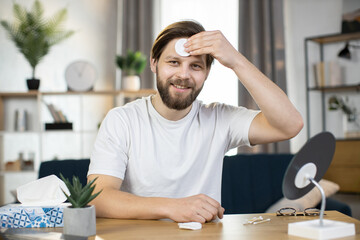 Attractive smiling Caucasian guy performs hygienic procedures, cleaning his face with cotton pad,...