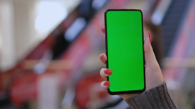 Hands of a young woman holding a smartphone in a shopping mall. A young woman is holding a phone in a shopping mall in front of an escalator with people. Close up