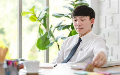 Portrait studio shot of Asian professional successful male businessman employee in formal shirt with necktie sitting look at camera at working desk with computer monitor keyboard mouse and stationery