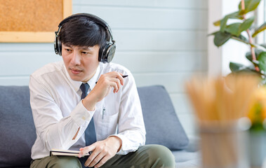 Asian young handsome professional successful male businessman employee in formal business shirt and necktie sitting on cozy sofa listening to streaming music online browsing internet with smartphone