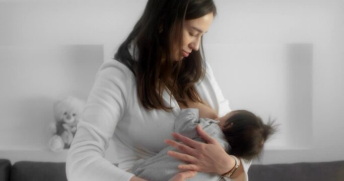 Mother is breastfeeding her newborn baby in the nursery. Mom in a white coat holds and cradles a infant in her arms. Smart watches on the hands of a modern mom. Concept of childhood, parenthood.