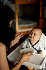 mom feeds the baby with a spoon . they are sitting at home in the kitchen and the light shines beautifully on them