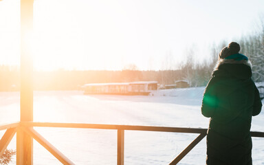 Back view of lonely young woman in warm clothes and hat standing on veranda and enjoying winter...