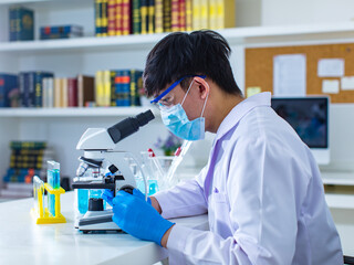 Asian professional male scientist in white lab coat and safety surgery face mask sitting typing keyboard computer analyzing experiment data information result on working desk in hospital laboratory