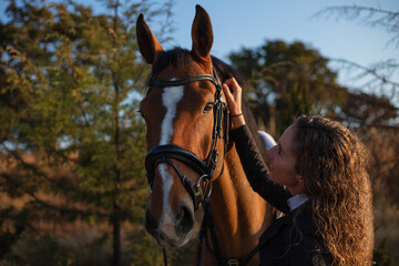 Young woman with curly hair petting her horse in a sunny morning