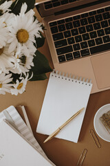 Laptop, spiral flip notebook, chamomile flowers bouquet on neutral brown background. Flat lay, top view aesthetic minimal freelancer, blogger home office workspace