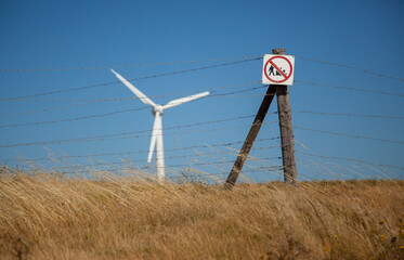 A wind turbine in a field with a wire fence in the foreground, with a sign prohbiting making a fire.