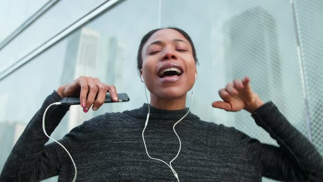 Happy african woman listening to music on mobile phone singing in the urban city street charming black young woman enjoy music dancing in the city