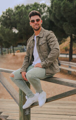 Obraz premium full length image of an attractive young man with sunglasses smiling and looking at the camera while is sitting on a wood railing of a viewpoint with the highway, mountain and trees in the background