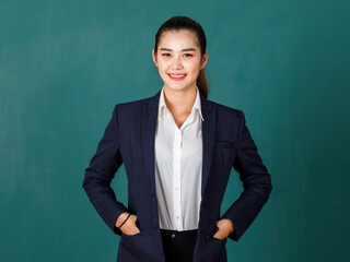 Portrait studio shot of Asian professional ponytail hair female teacher or college student in formal suit standing smiling look at camera hold hands in jacket pockets on green chalkboard background