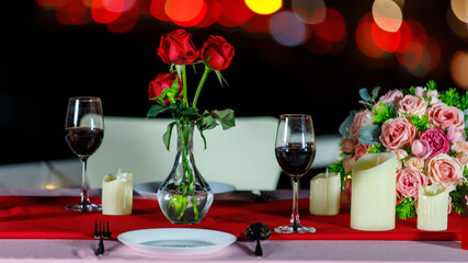 Closeup shot of full decorated fine dining table with tablecloth full of dinner equipment wine glasses fork spoon dishes candles red roses vase and bouquet on bokeh background in open air restaurant