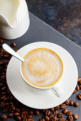Coffee and coffee beans on a slate board. Cup of coffee on a white saucer and a milk jug on a dark background