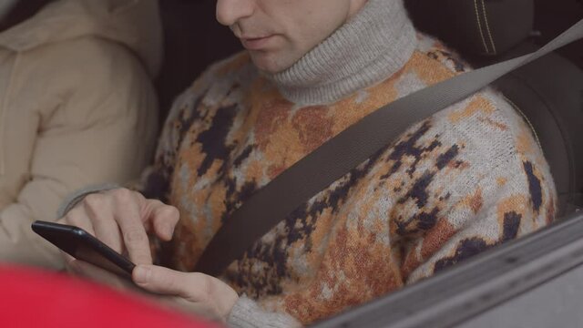 Tilting slowmo shot of Caucasian man with fastened seat belt scrolling on smartphone sitting at driver seat in car with his girlfriend