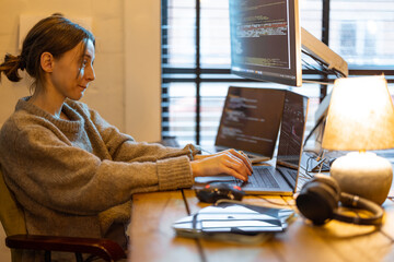 Young focused woman writing programming code, sitting at workplace with laptop and desktop...