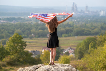 Young happy american woman with long hair raising up waving on wind USA national flag in her hands...