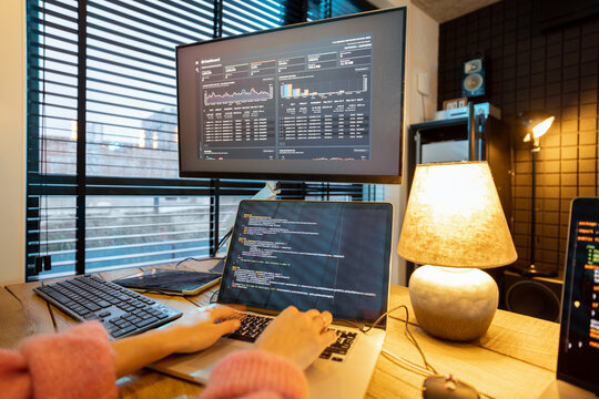 Female programmer writing programming code on laptops and desktop computer at cozy home workplace. Close-up on hands and keyboard
