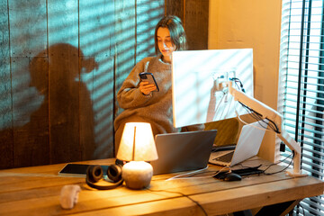 Young focused woman using smart phone while standing at cozy home office during the nighttime. Concept of online work from home. Idea of cozy and style workplace