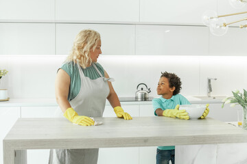 Caring mom teaching her son how to clean table with detergent in white kitchen