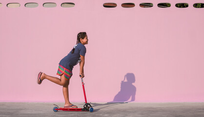 Asian little dark skinned girl has fun by riding kick scooter near pink wall on the patio outside...