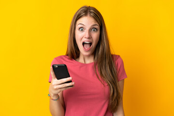 Young woman using mobile phone isolated on yellow background with surprise and shocked facial...