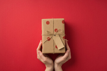 First person top view photo of valentine's day decorations woman's hands demonstrating craft paper giftbox with polka dot pattern and twine bow on isolated red background with empty space