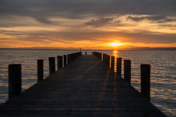 Atmospheric photo of the sunset at Lake Neusiedl with a wooden pier in the foreground. The wooden poles cast a shadow on the jetty. The orange cloudy sky with the turquoise wavy water