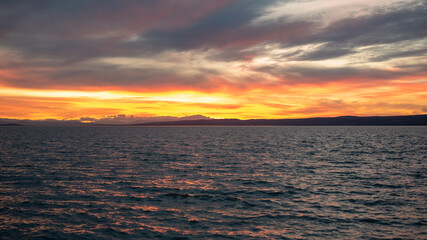 Twilight at Lake Neusiedl with sun reflections in the water, with cloudy orange-red blue sky. In the background the silhouette of the mountains and rippled water. Calming mood.