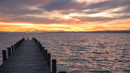 Dusk at Lake Neusiedl with a wooden pier in the left foreground. The turquoise rippled water with an orange reflection streak in the background. The orange red cloudy sky 