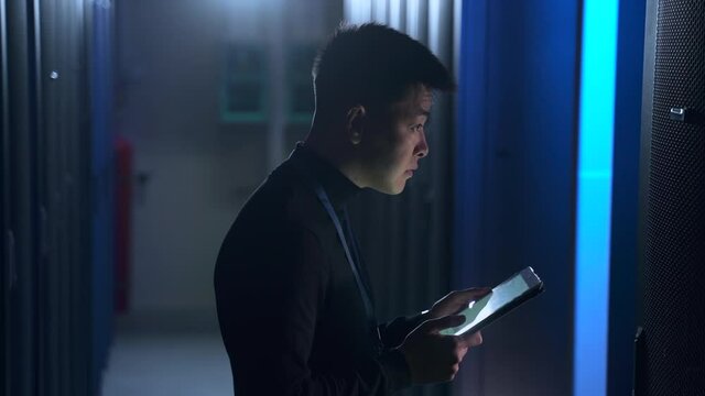 Young businessman or engineer using tablet and checking hardware, standing in data office spbas. Side view of Asian man holds device in hand and examines equipment, types report and stands in dark