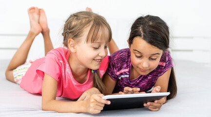Two cute little girls are using a digital tablet and smiling while lying on bed in children's room