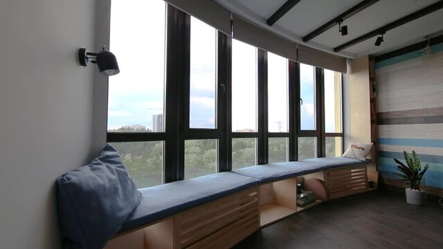 Video of motorized roller shades in the interior. Automatic roller blinds beige color close down on big glass windows. Remote Control Shades are above the windowsill with pillows. Summer. 
