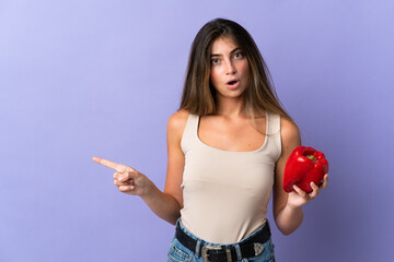 Young woman holding a pepper isolated on purple background surprised and pointing side