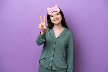 Young Russian woman in pajamas isolated on purple background smiling and showing victory sign